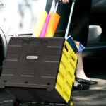 A person sporting black shoes and pants is pulling a yellow foldable rolling cart labeled "#2198 Foldable Rolling Cart w/ Lid Cover Yellow 16" X 18" X 15"." The cart, which also features black accents, is brimming with colorful rolled-up papers and various supplies. The person stands beside an open car door.